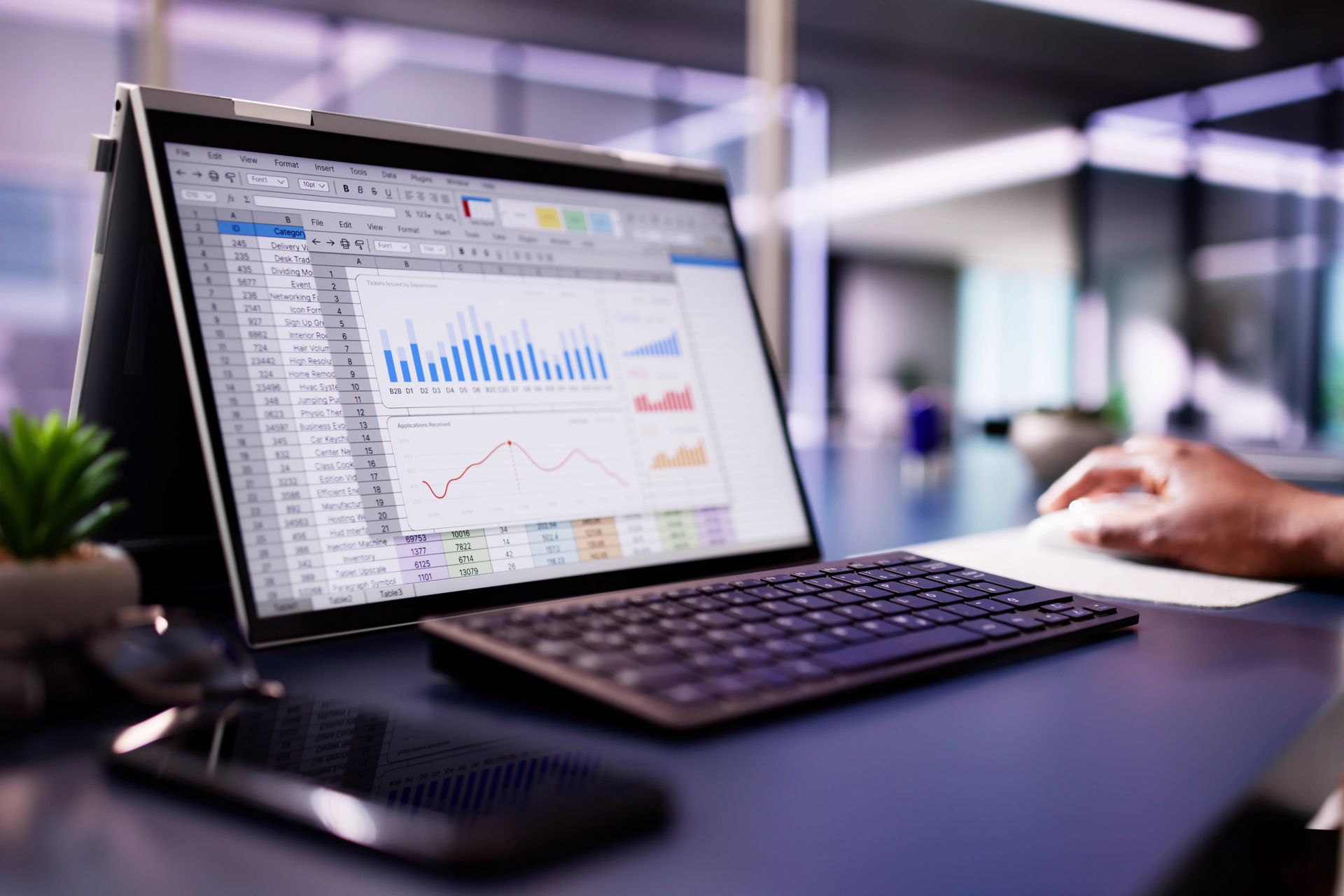 Laptop displaying graphs and data on a desk, with a keyboard, phone, and hand.