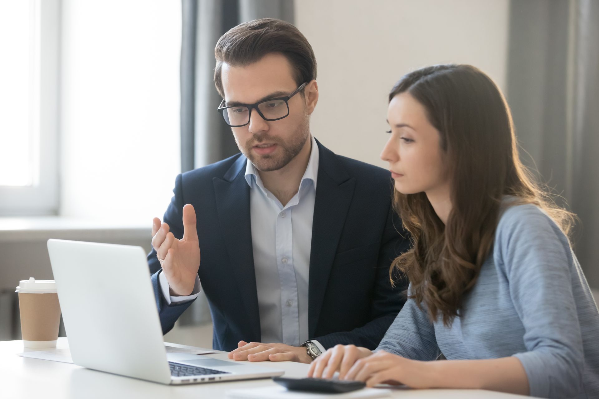 Man in suit gestures at laptop, explaining to woman in blue, both in an office setting.