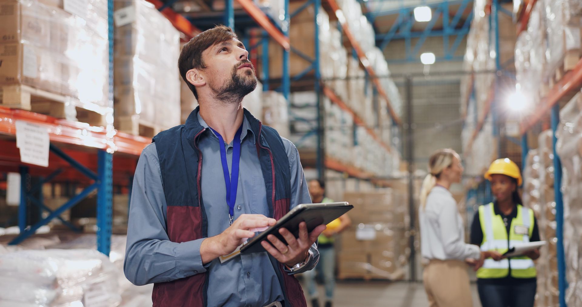 Man in warehouse, using tablet, looking up. Others in background, shelving, boxes.
