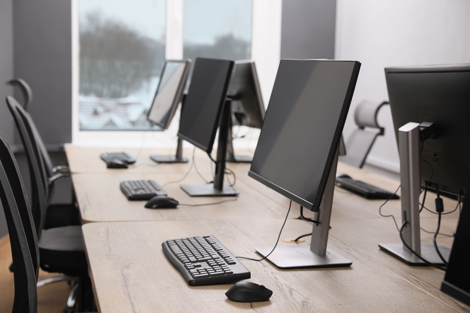 Computer workstations in an office. Monitors, keyboards, and mice on a light wood desk, chairs lined up. 