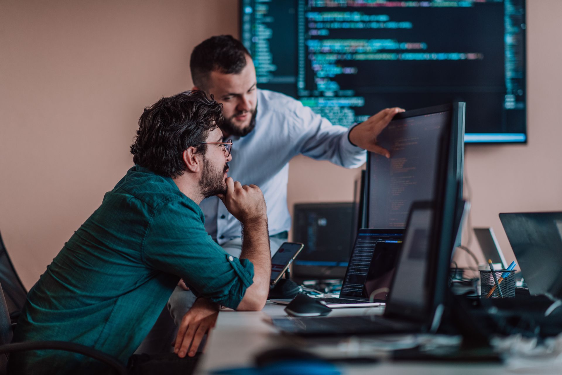 Two men collaborating at computers, pointing at code on screen in office.