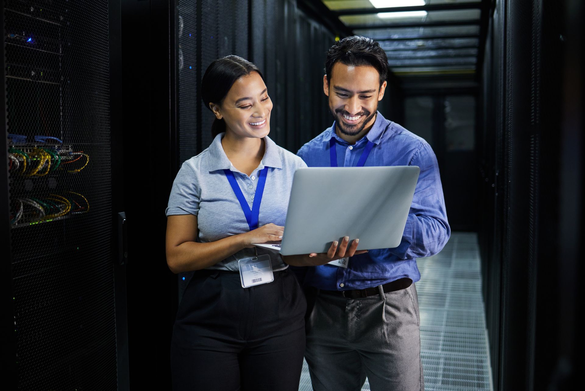 Two people in a server room looking at a laptop, smiling.