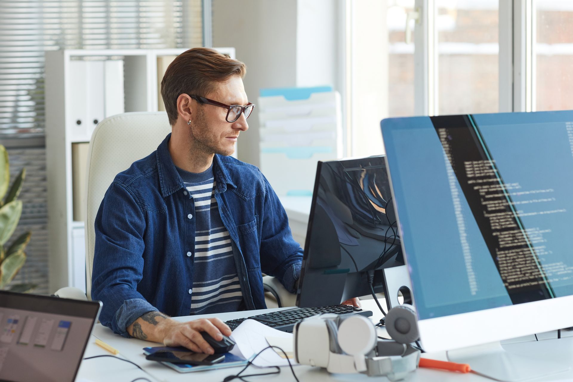 Man with glasses working on computers in an office. Displays show code. He wears a denim shirt.
