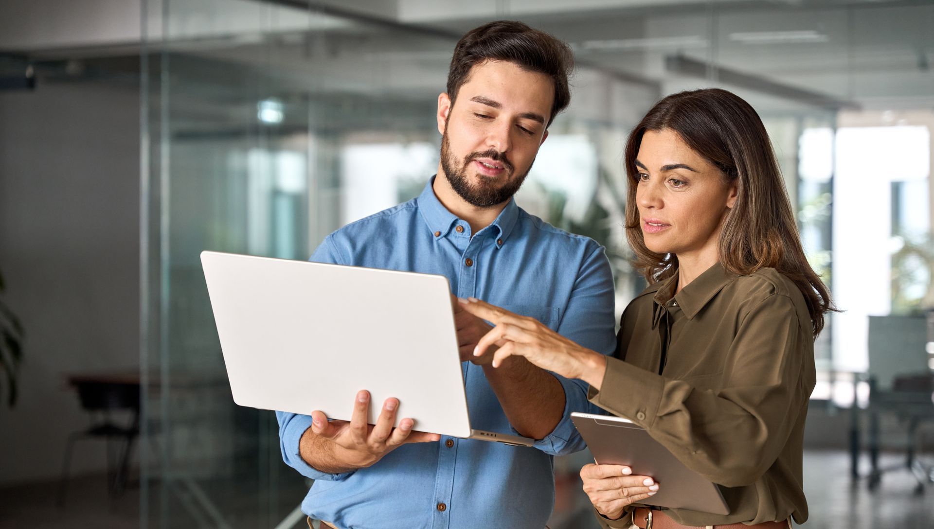 Man and woman in office looking at laptop, woman pointing at screen, holding a notepad.