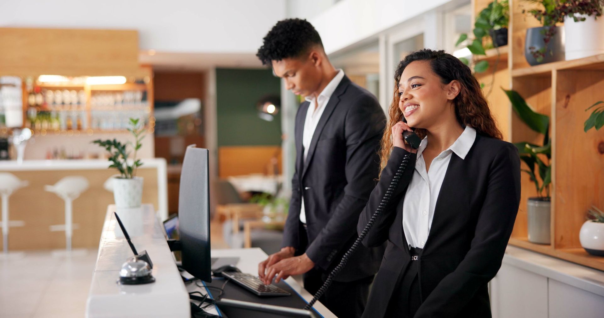 Two hotel staff members at a reception desk; one on the phone smiling, one typing on a keyboard.