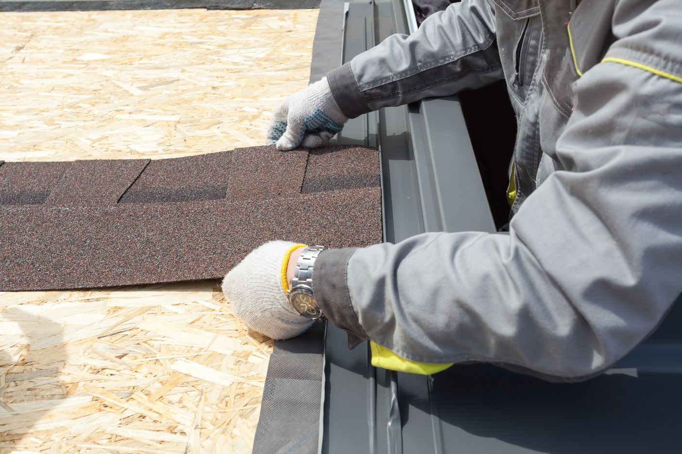 Worker installing brown roofing shingles on a roof edge with gray gloves and a metal trim piece