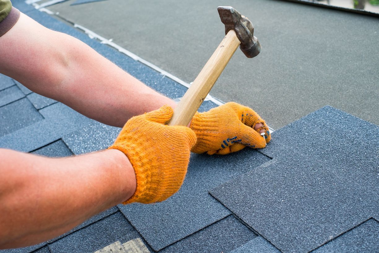 Gloved hands using a hammer to nail blue shingles on a roof