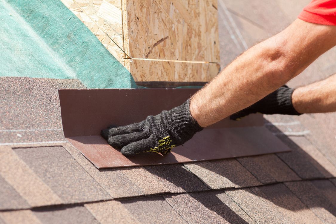 Gloved hand installing a maroon roof shingle on a sloped roof near a chimney base