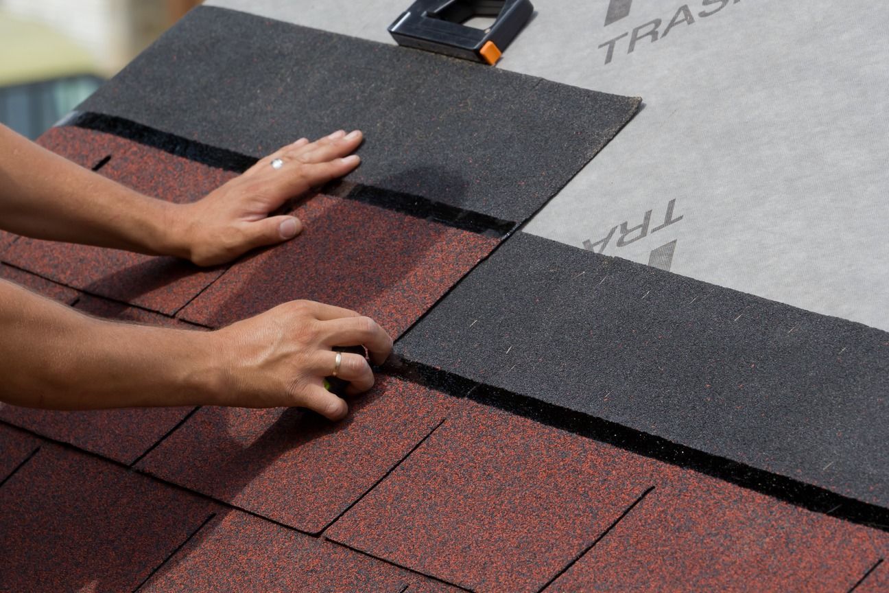 Hands installing dark shingles on a roof with red underlayment visible