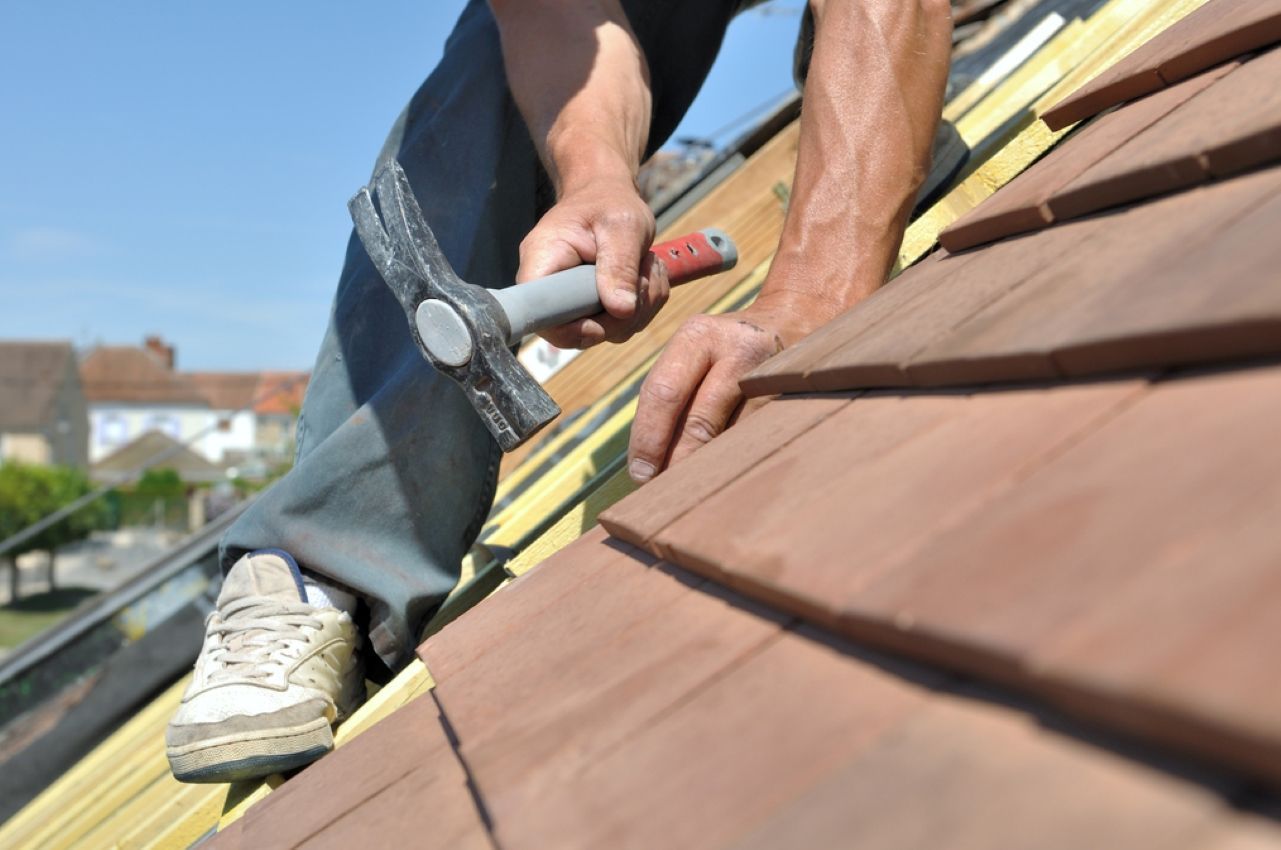Roofer hammering nails into brown roof shingles while kneeling on a sloped roof
