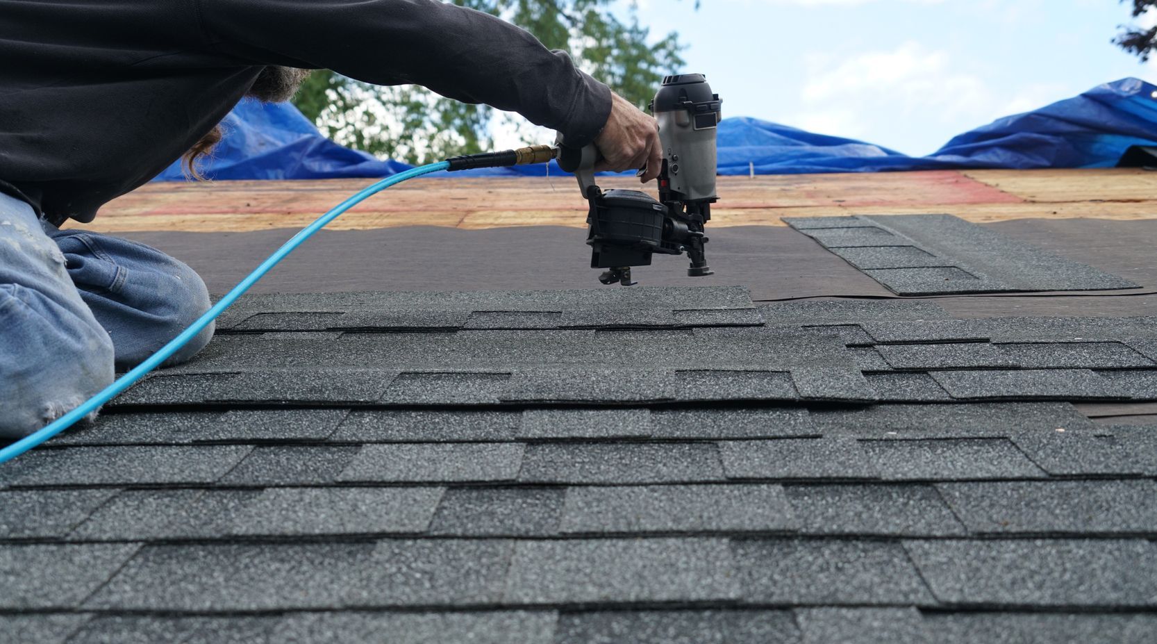 Worker using a nail gun on a shingled roof, with blue tarp and safety rope visible.