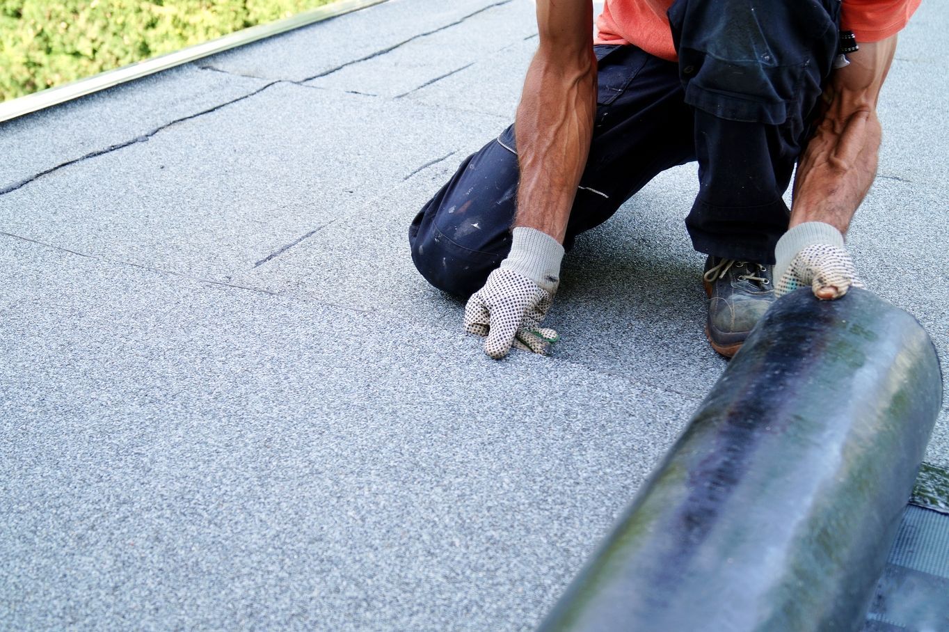 Worker kneeling on a roof, rolling black waterproof roofing material over a gray surface.