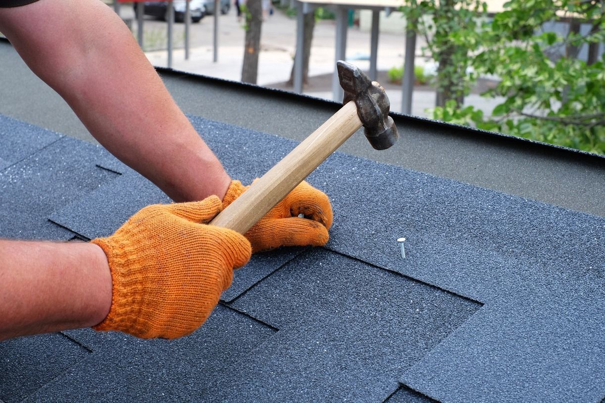 Hands in orange gloves using a hammer to repair blue roof shingles