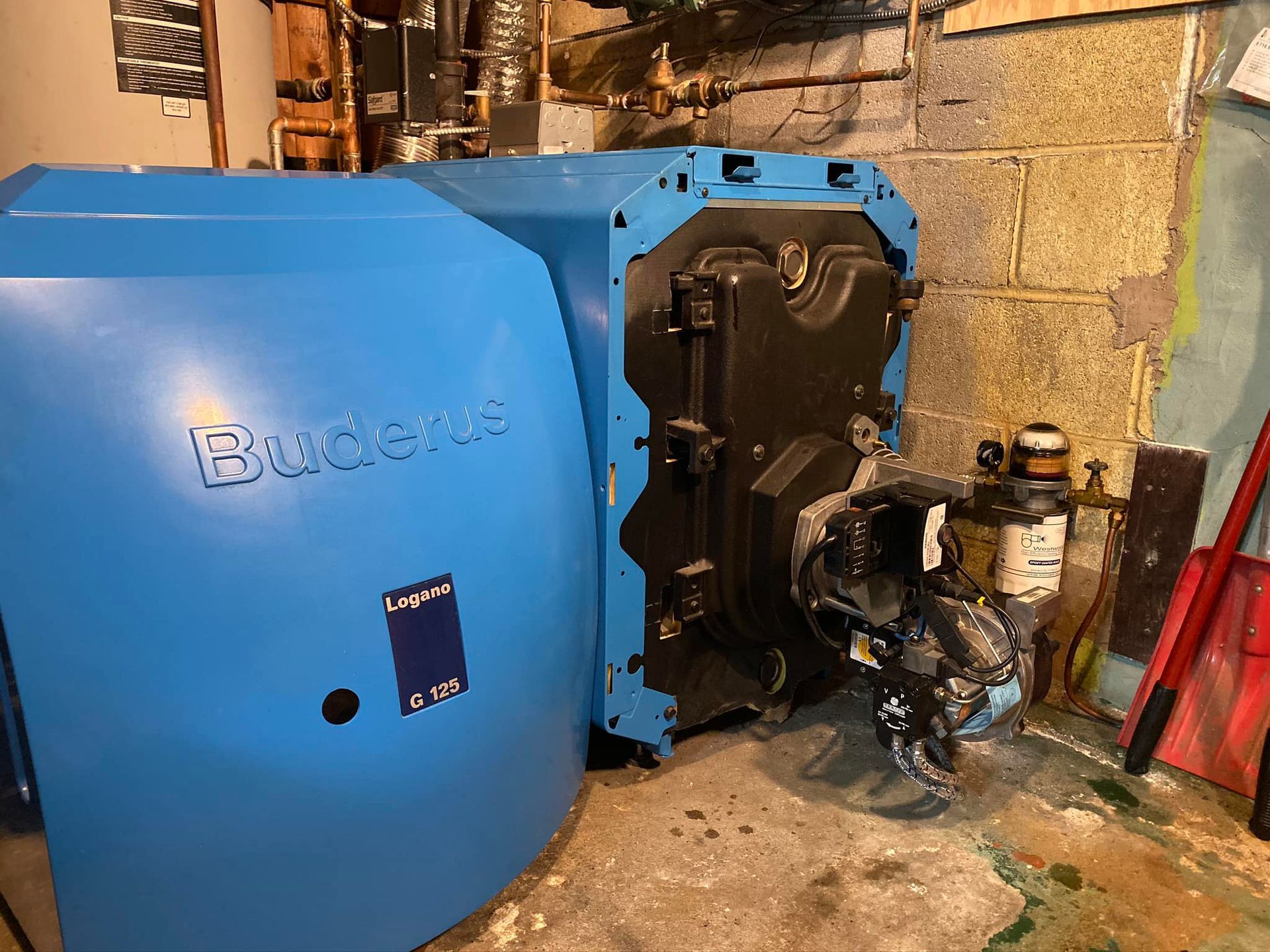 A mechanical room showing a cylindrical HTP water heater connected by copper pipes to a metal boiler against a block wall.