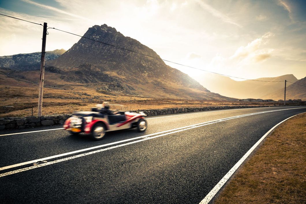 Classic car speeding along a scenic road with mountain backdrop and bright sunset.