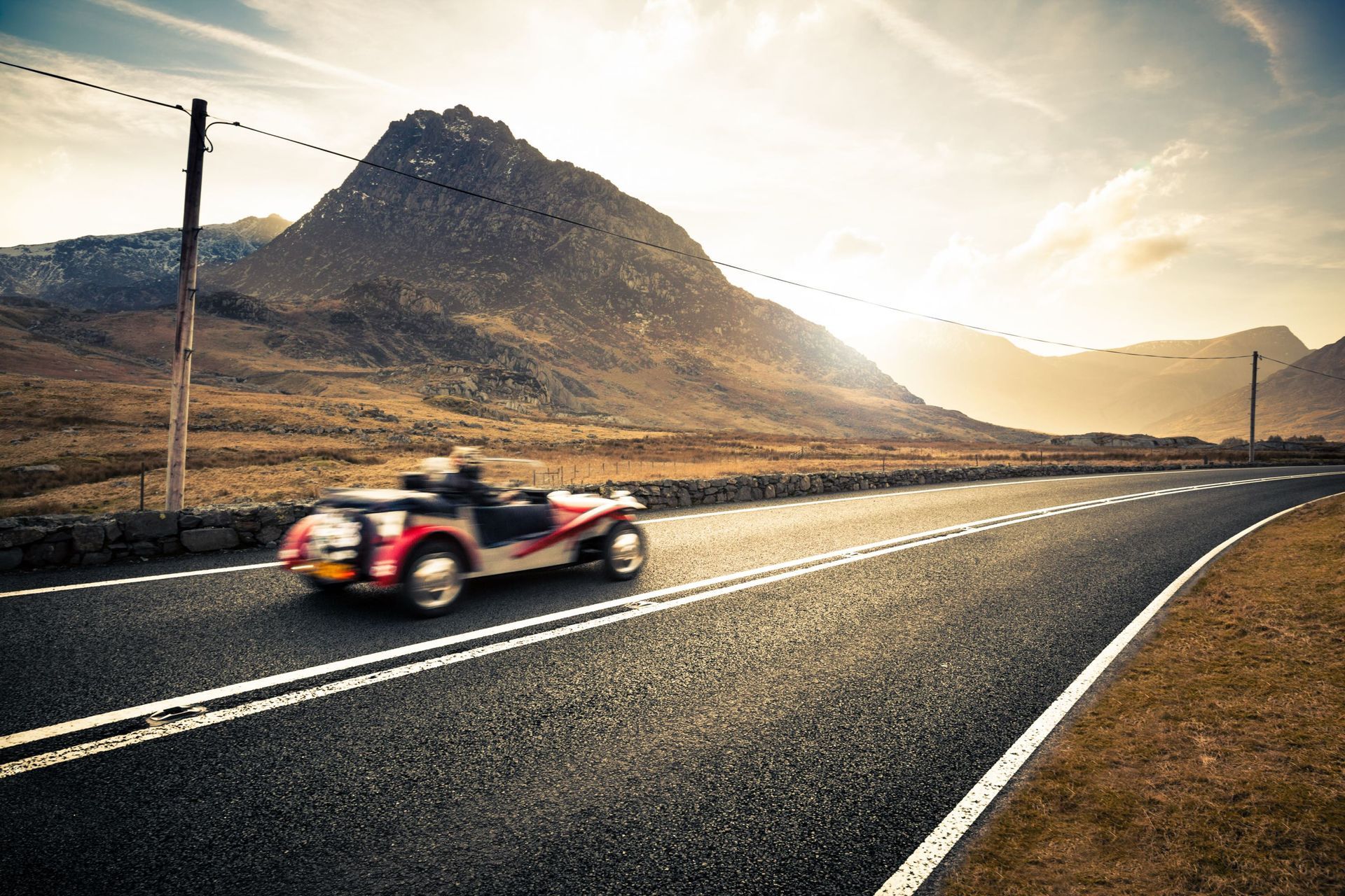 Classic car speeding along a scenic road with mountain backdrop and bright sunset.