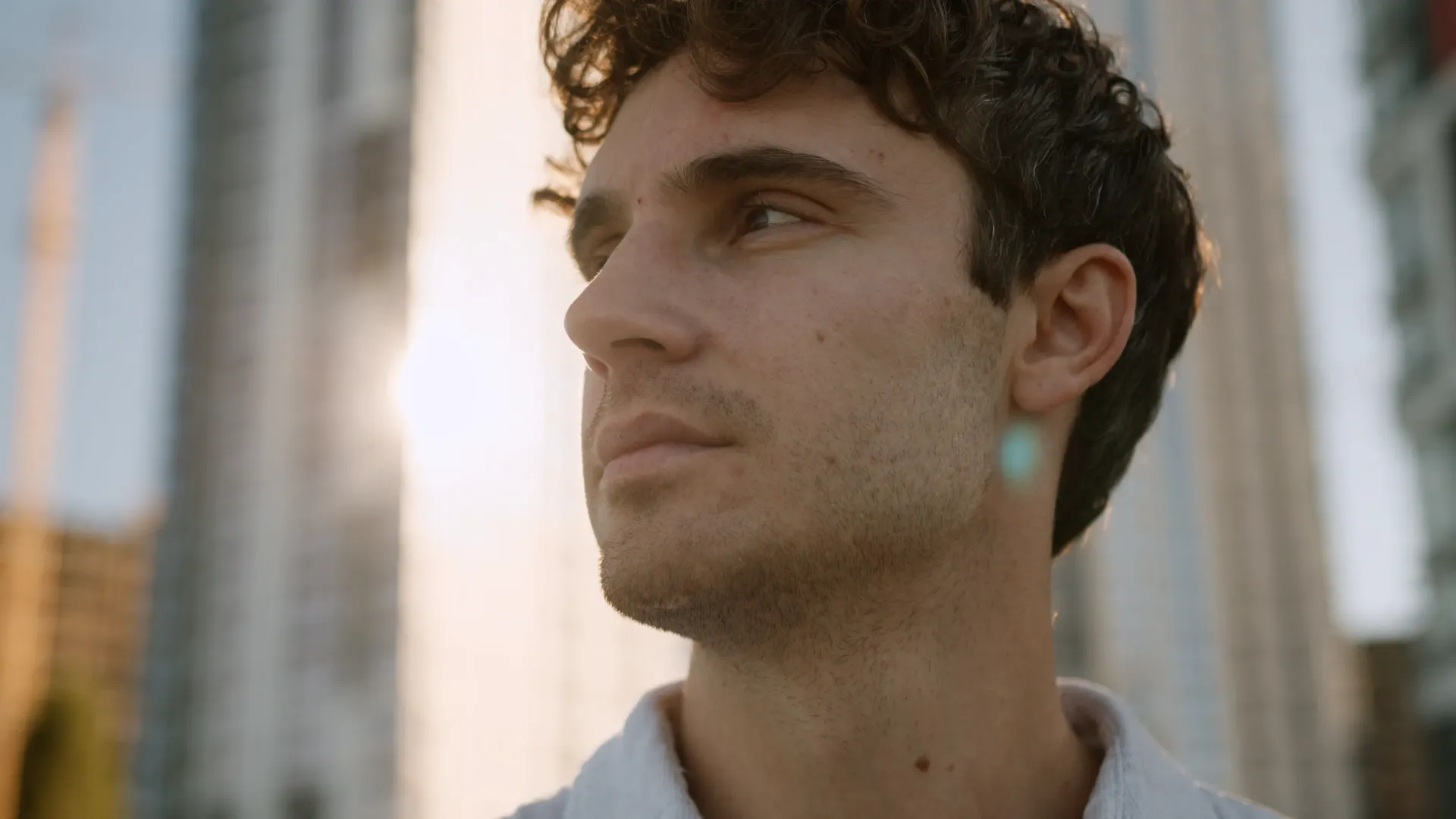 Man with curly brown hair looks off to the side in front of tall buildings, bathed in sunlight.