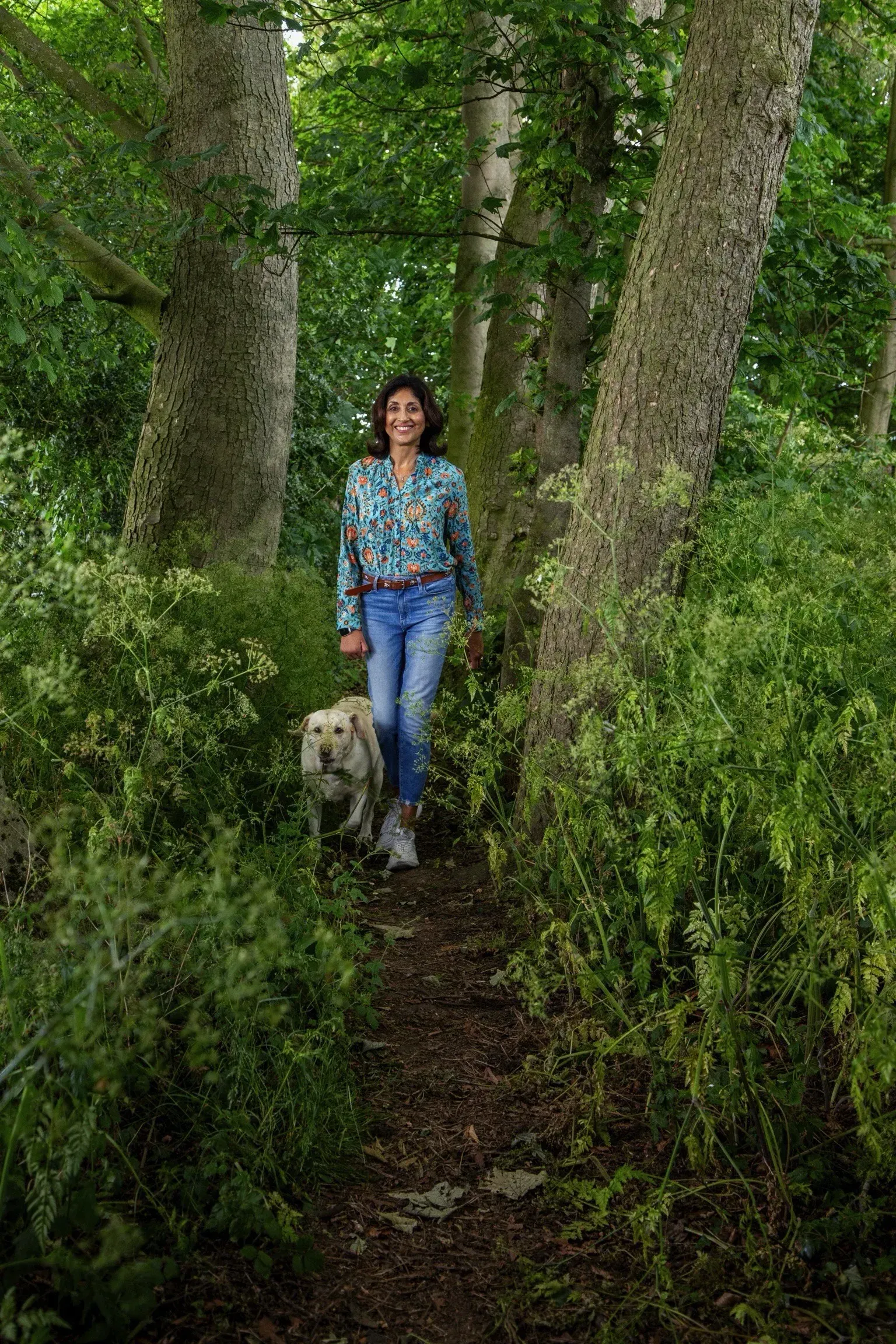 Satinder walking dog on a wooded path, smiling. She wears jeans and a patterned shirt, surrounded by greenery.