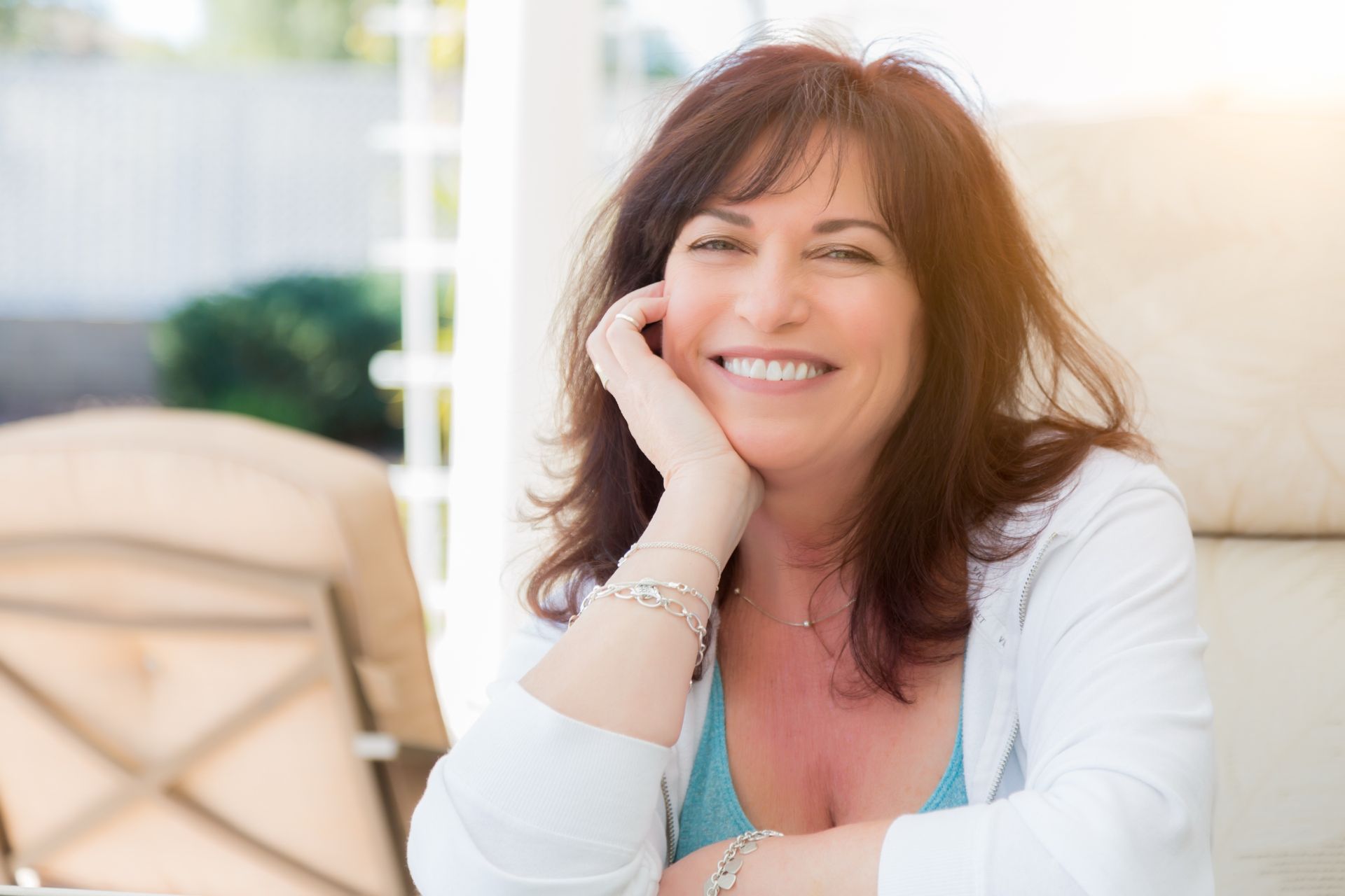 Woman with dark hair smiles, resting chin on hand, sitting outdoors.