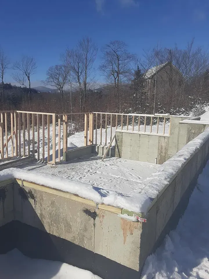 A building under construction in the snow with a house in the background.