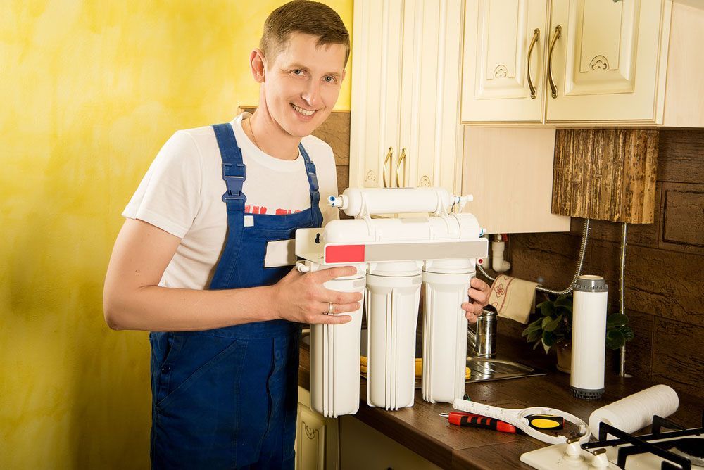 A Man Is Standing In A Kitchen Holding A Cup Of Water And A Water Filter — Eureka Water & Filtration In Mount St John, QLD