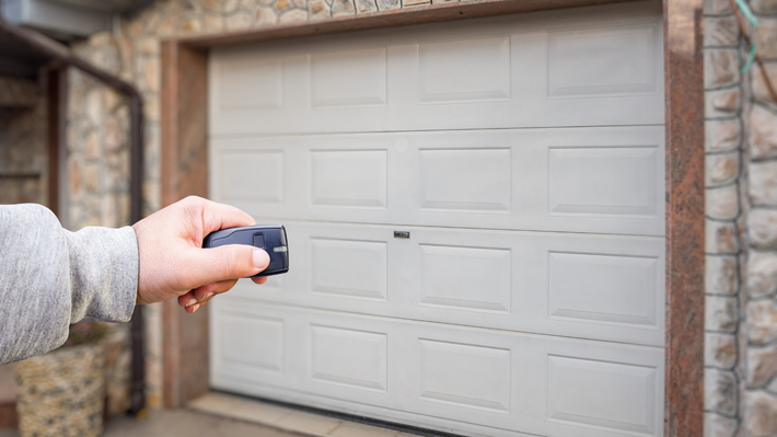 Hand holding a garage door opener, aiming at a closed white garage door in front of a stone wall.