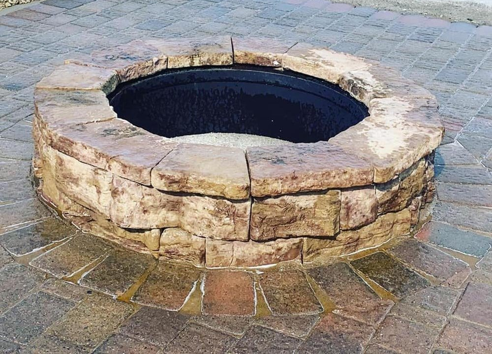 Backyard with a decorative block fire pit, brick paver patio, and house. A red car and American flag are in the distance. hardscape. fire features. wood burning. gas burning fire pit. 