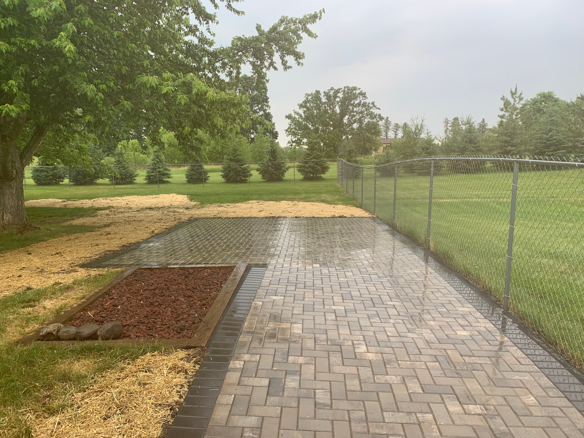 A brick walkway leading to a fenced in yard with trees in the background.