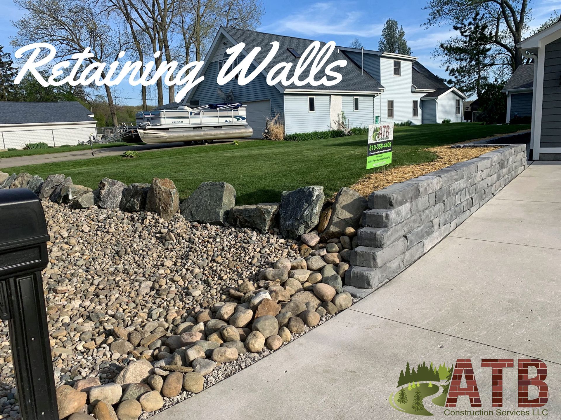Retaining wall made of grey blocks next to a boulder river rock garden and a driveway, with a house in the background. holding dirt back on hill. 