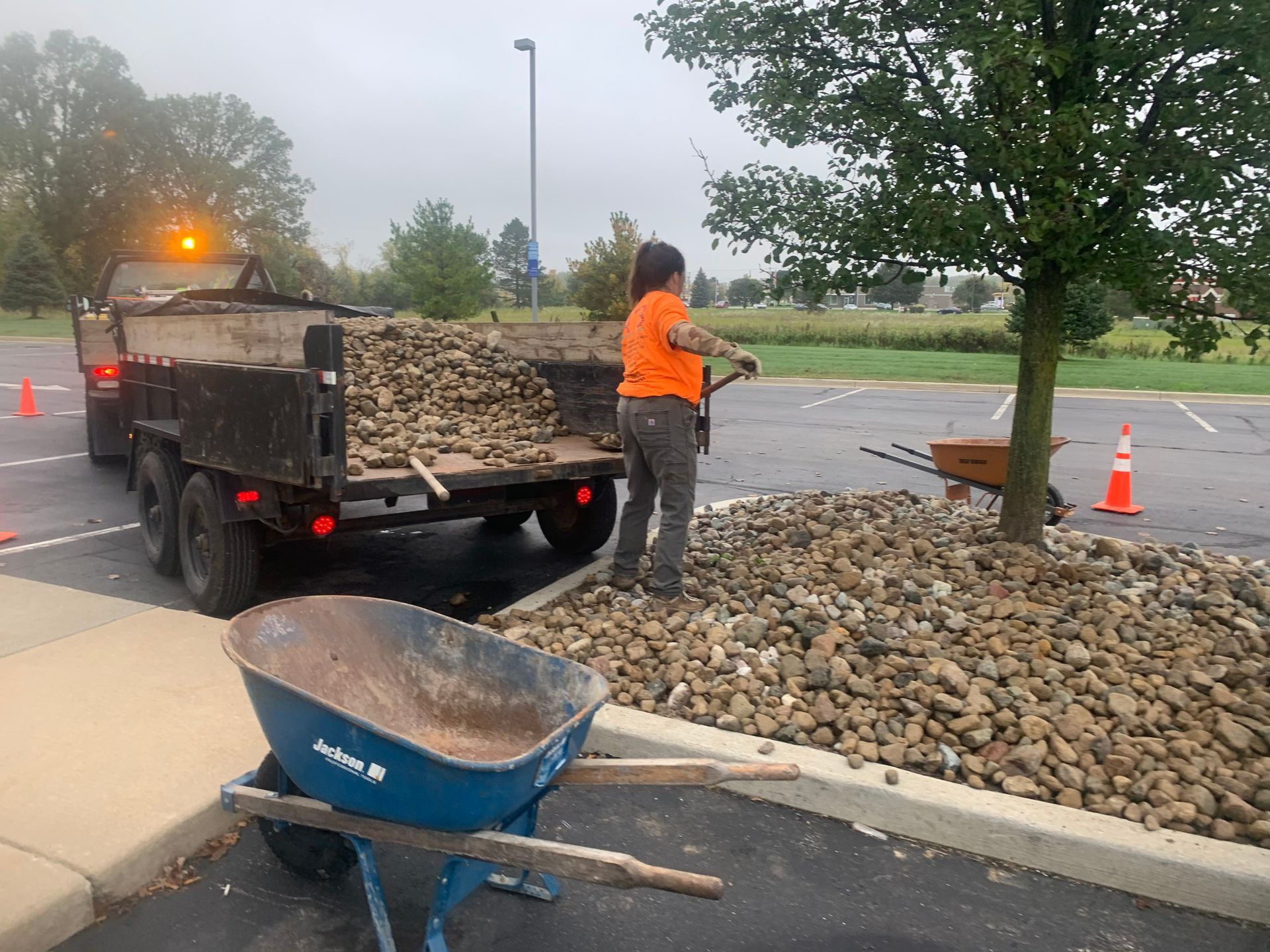 A woman is standing next to a wheelbarrow in front of a dump truck. mulch