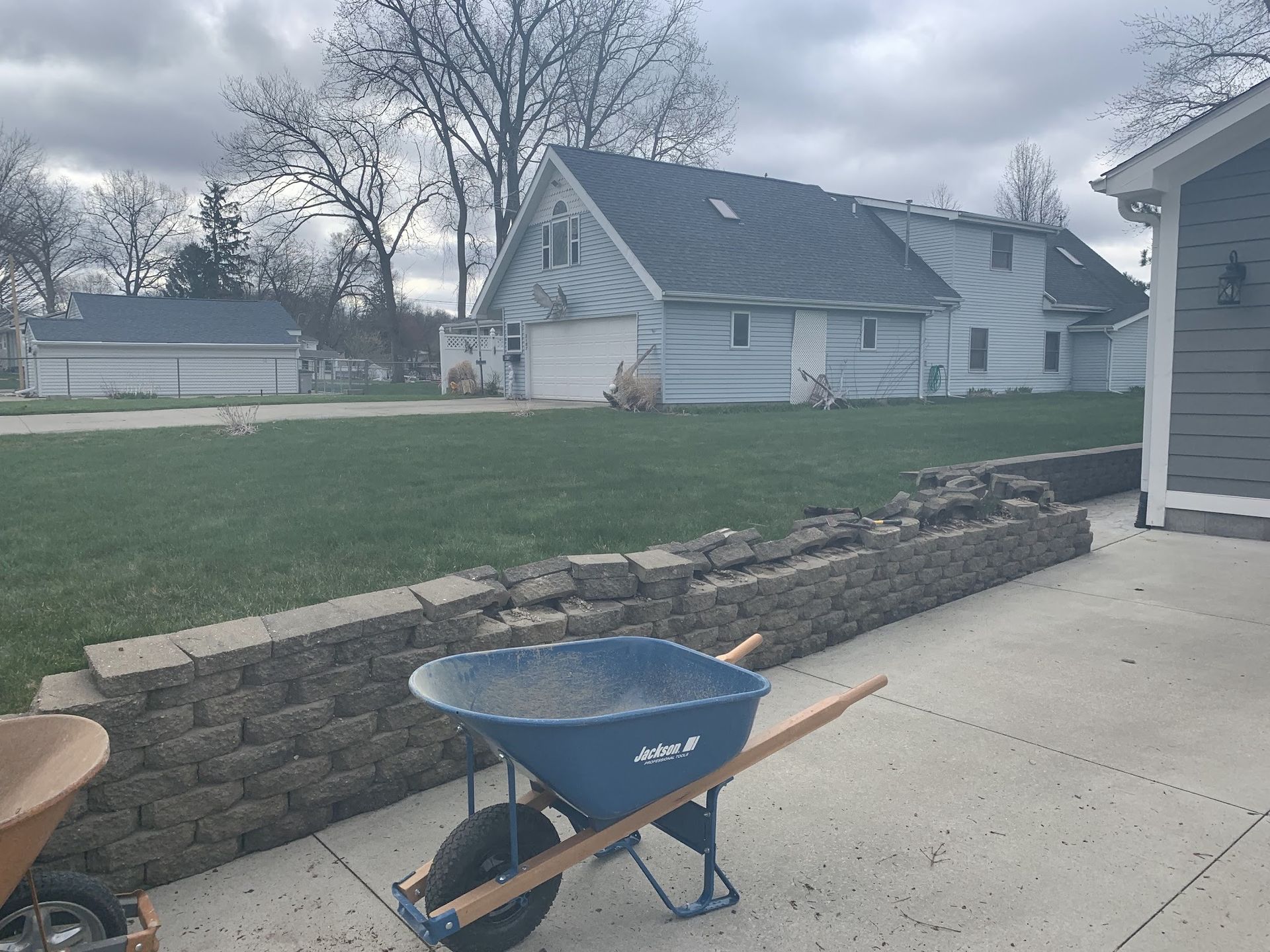 A blue wheelbarrow sits in front of a partially built retaining wall and a house with a garage.