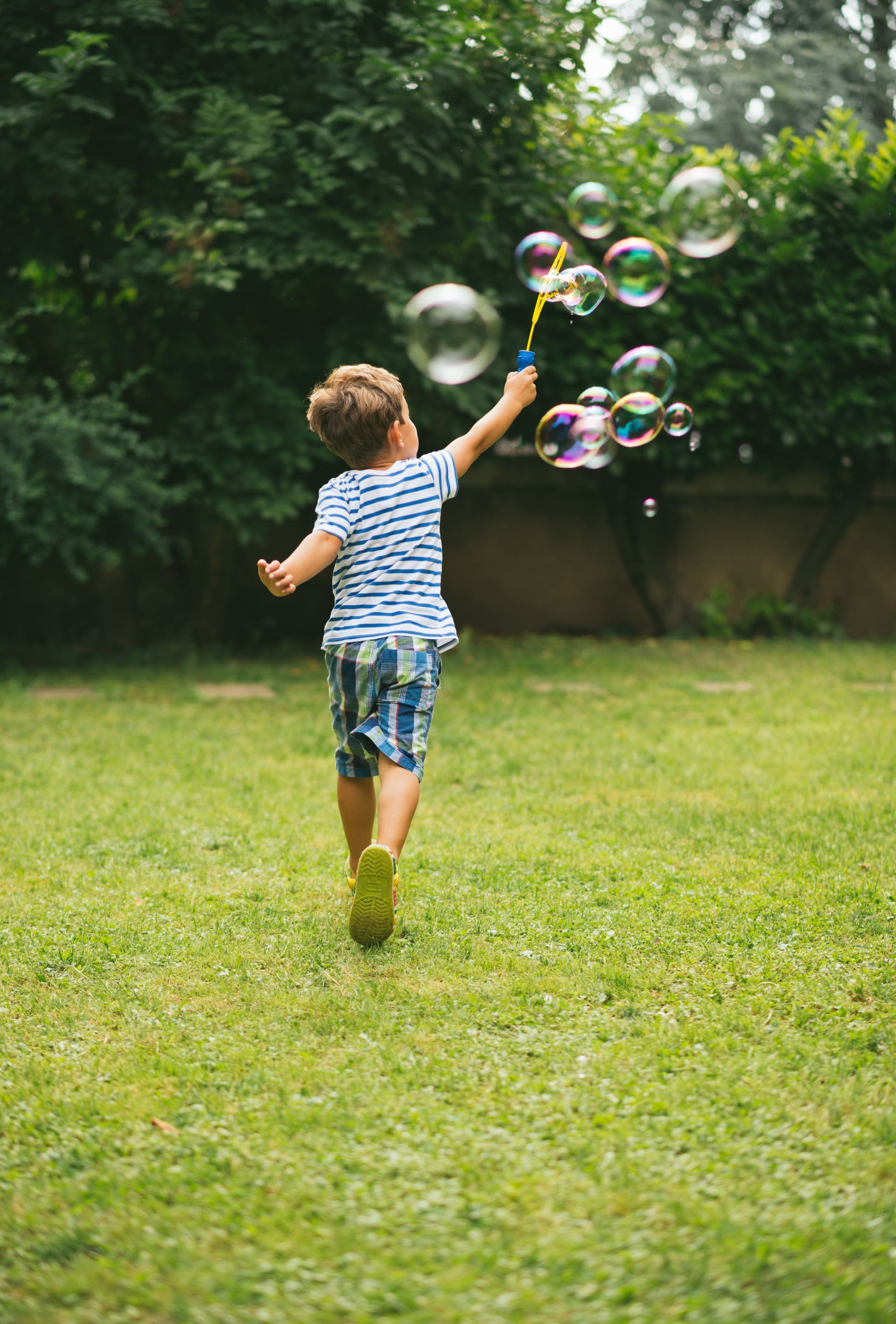 Child running on grass, blowing bubbles with wand in backyard.
