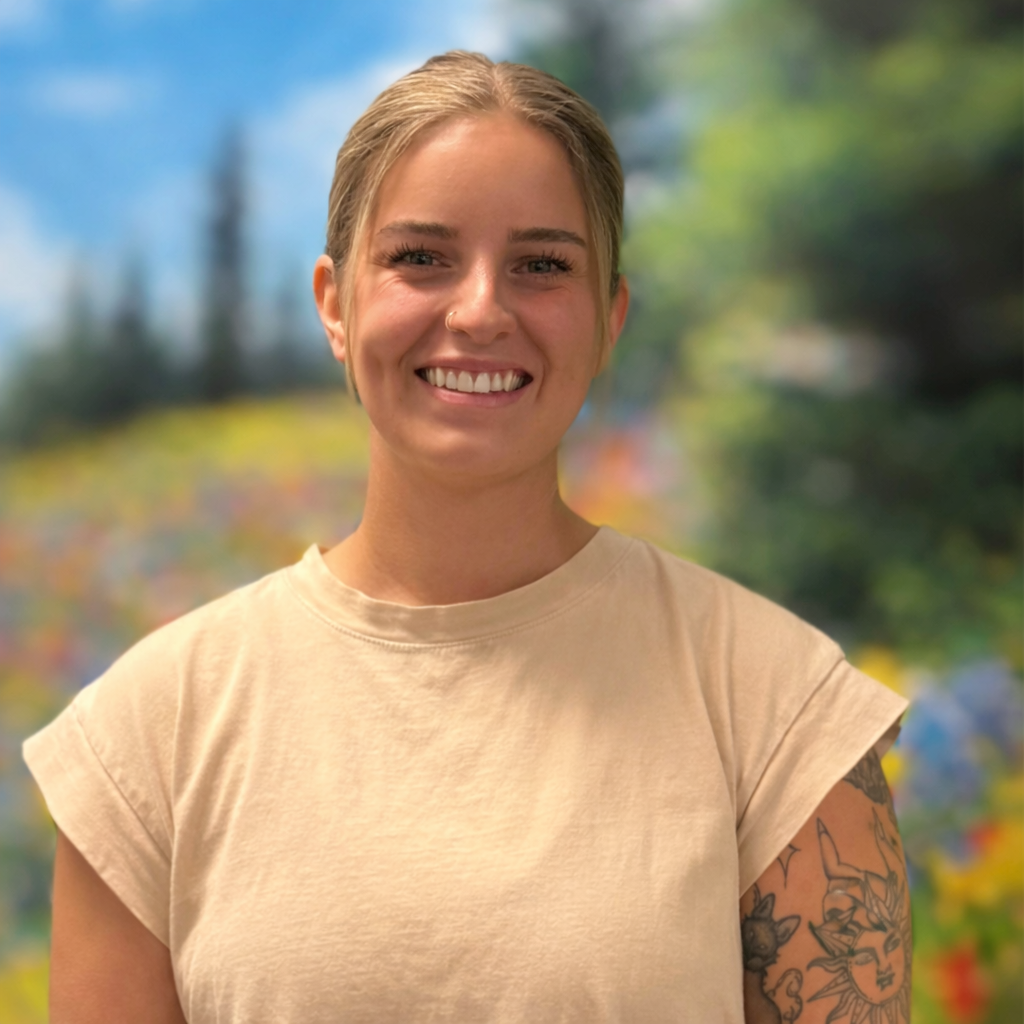 Woman with blonde hair smiles, standing in front of a floral backdrop; tattoo on arm.