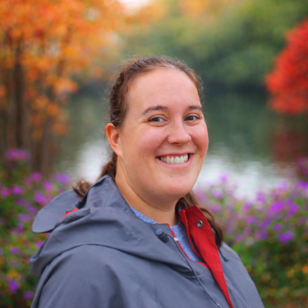 Woman smiling in front of fall foliage and water. She wears a gray hooded jacket and purple flowers are visible.