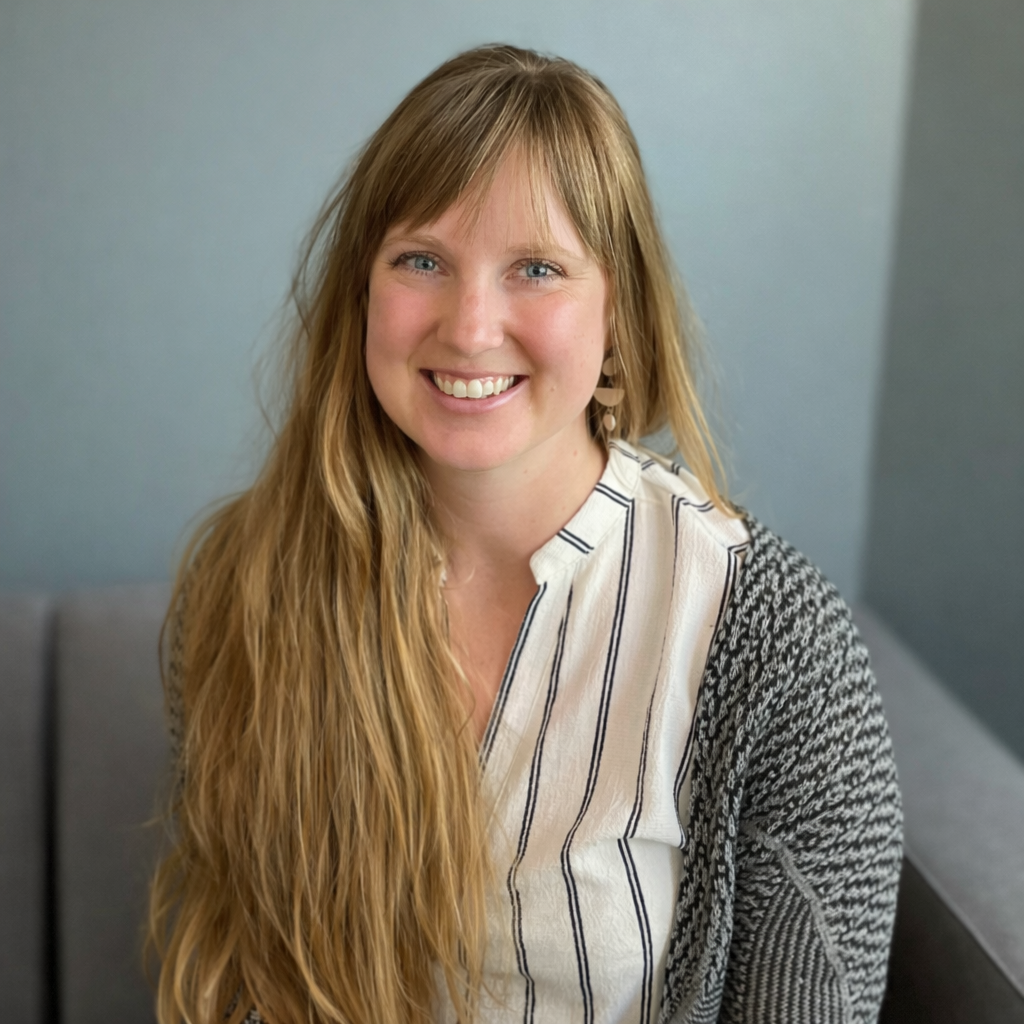 Woman with long blonde hair smiles, wearing a black and white patterned shirt, seated in a room with blue wall.