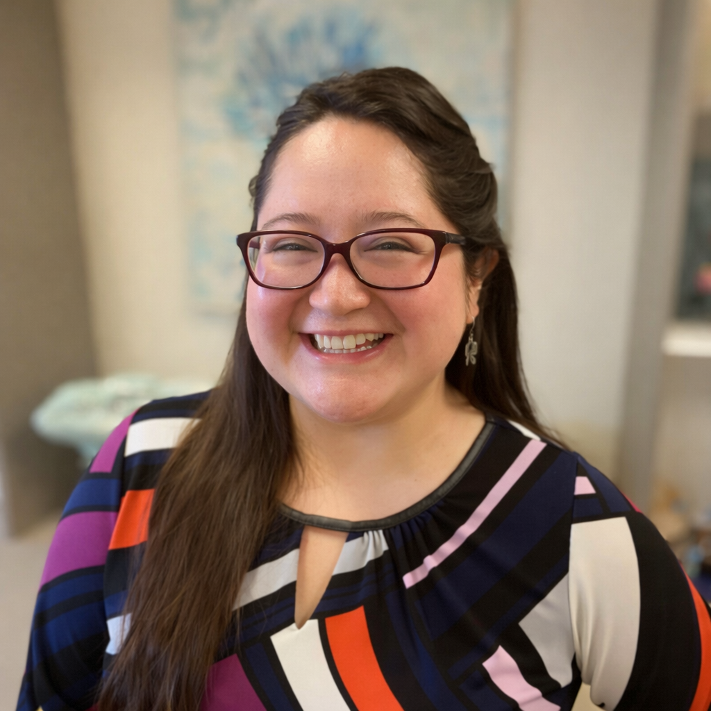 Woman in glasses smiles, wearing patterned top, stands in front of a blue and white painting.