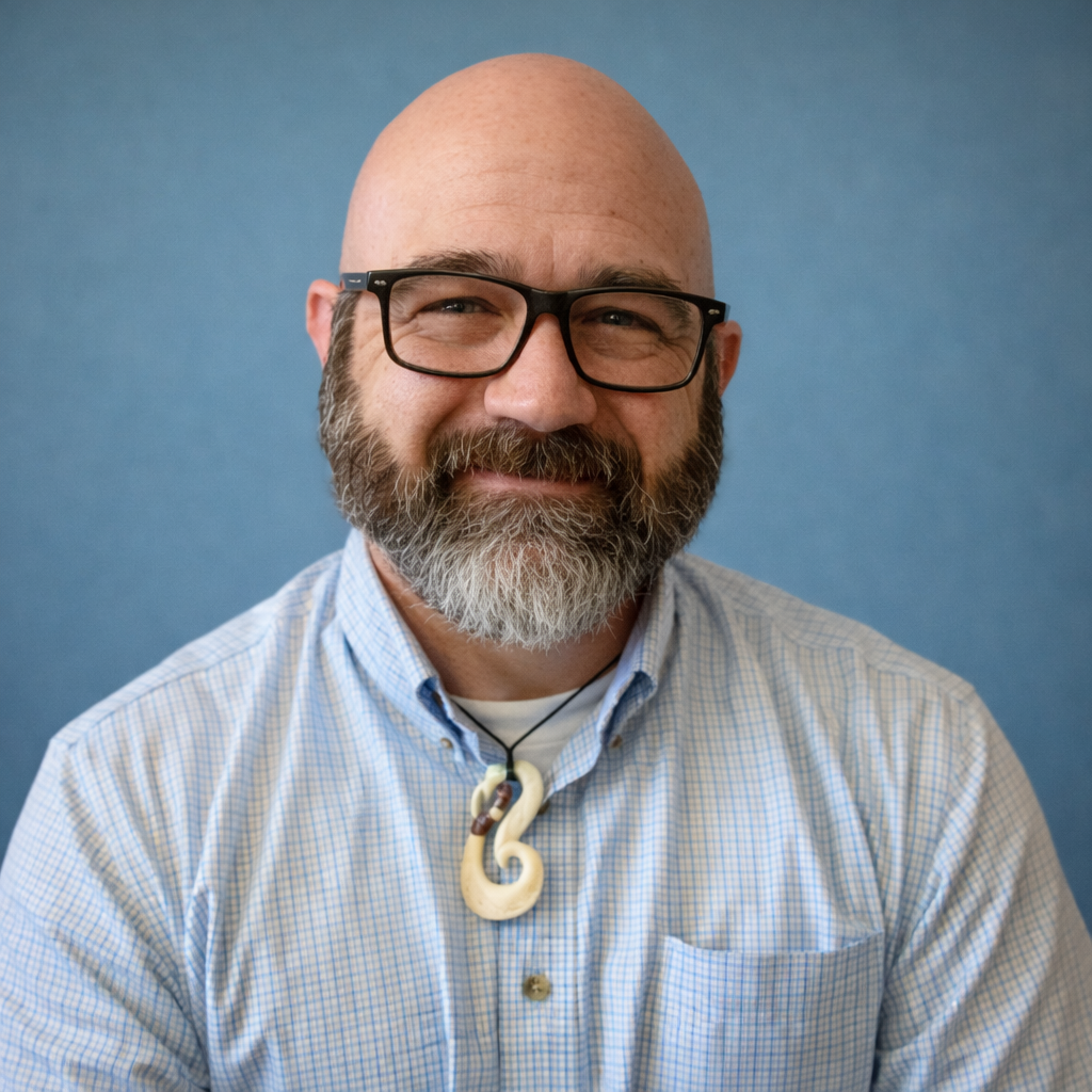 Man with glasses and beard wearing a checked shirt and bone pendant. Sitting indoors with patterned background.
