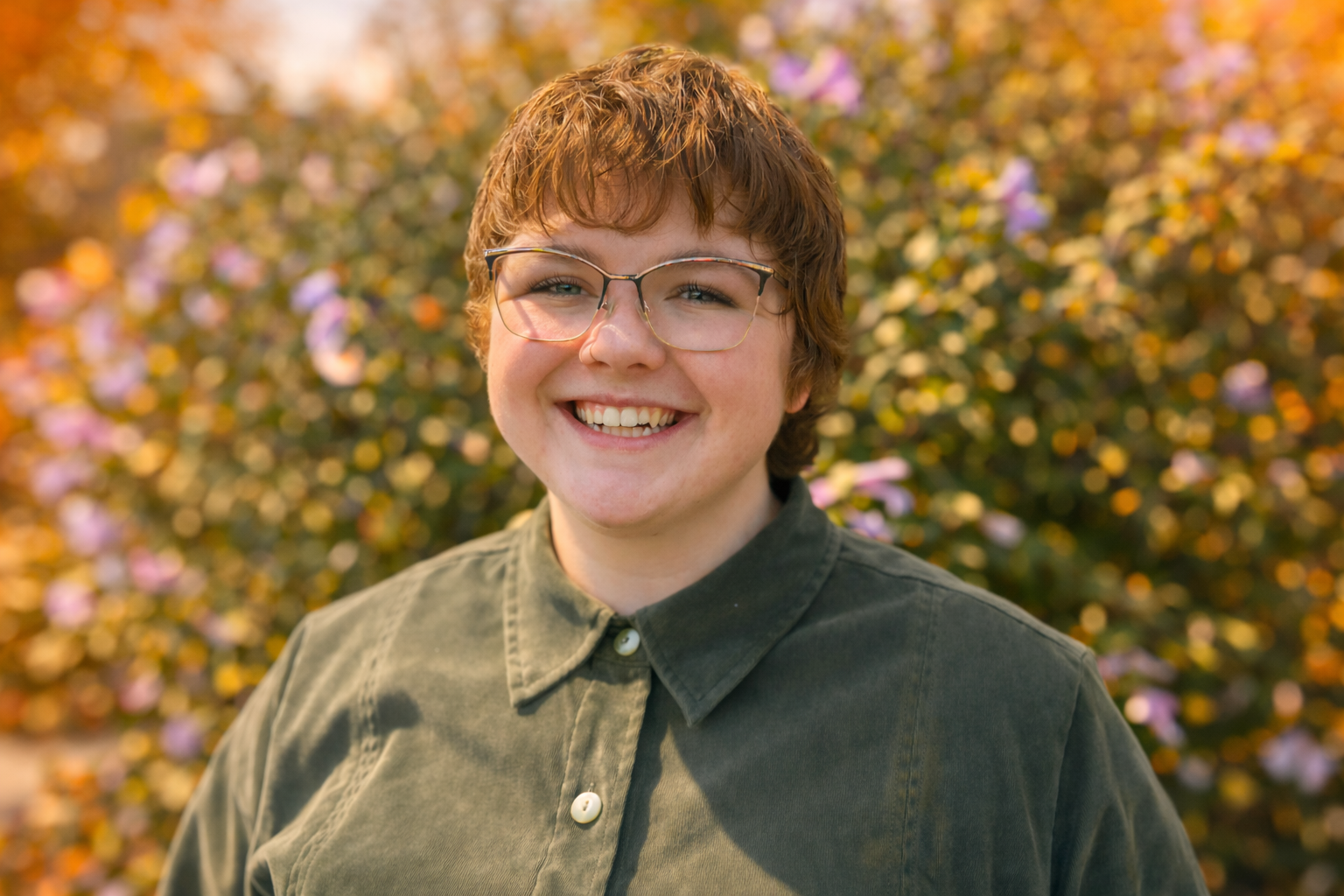 Person wearing glasses smiles in front of a bush with purple flowers; wearing a green collared shirt.