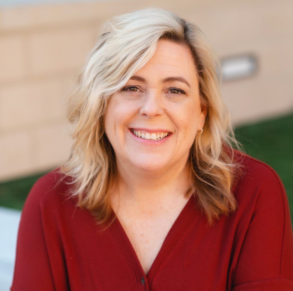Blond woman smiling, wearing a red sweater, outdoors in front of a blurred background.