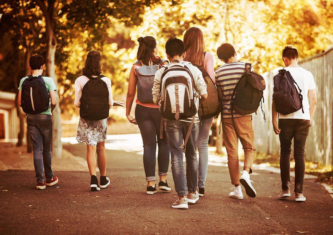 Group of students walking away on a sidewalk with backpacks, trees in the background.