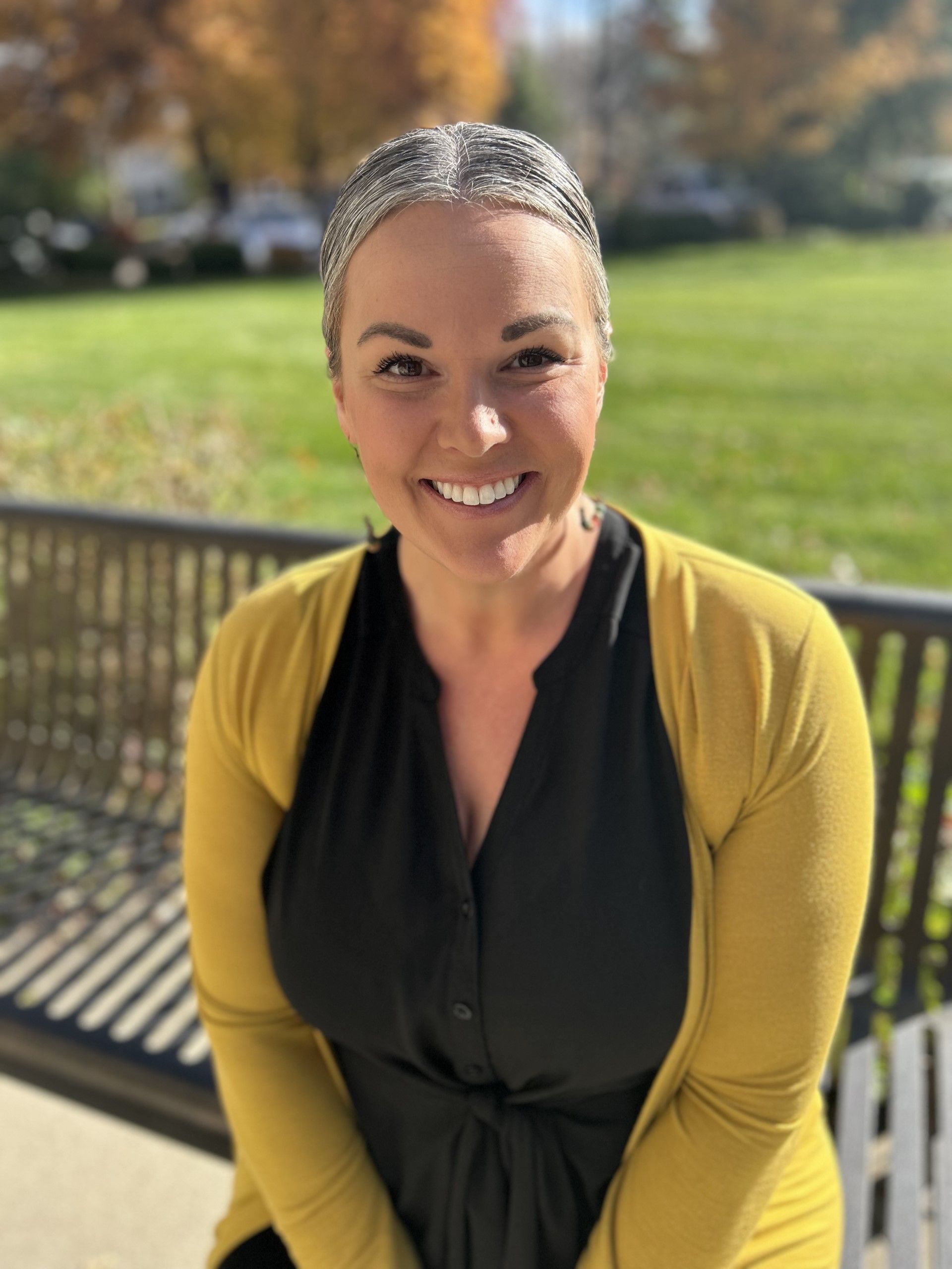 A woman wearing a yellow cardigan and a black shirt is sitting on a park bench.