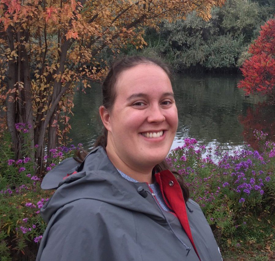 A woman in a grey jacket is smiling in front of a lake