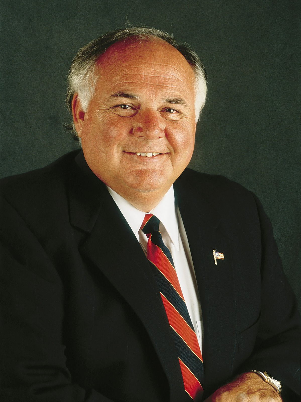 Man in a suit smiles at the camera, red and black striped tie, dark background.