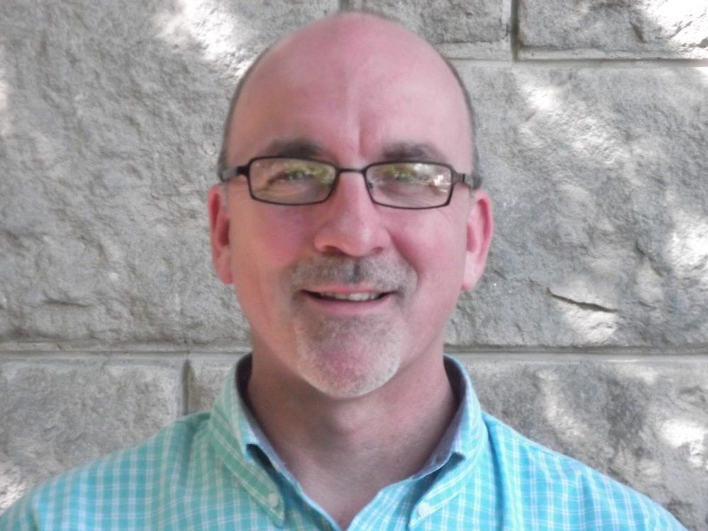 A man wearing glasses and a blue shirt is smiling in front of a brick wall.