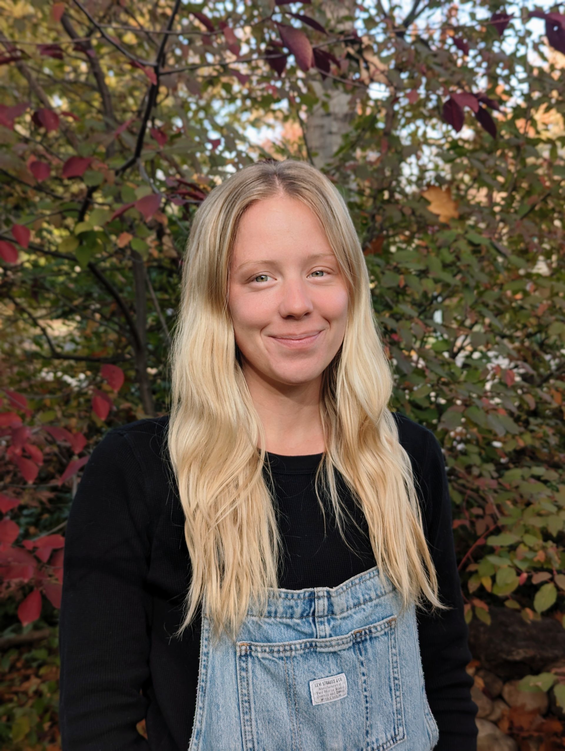 A woman wearing overalls and a black shirt is standing in front of a tree.