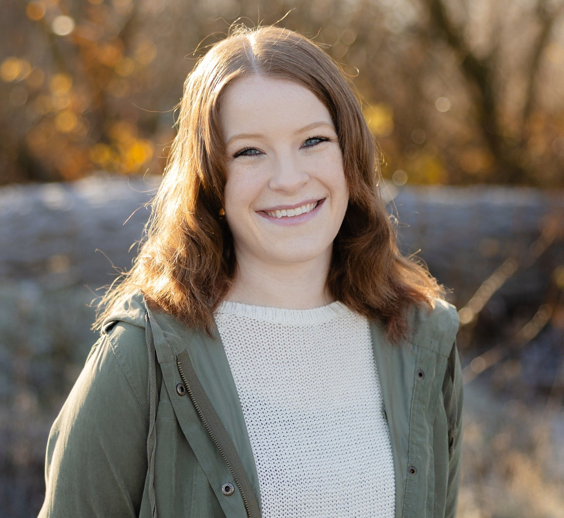 Woman with auburn hair smiles, wearing striped shirt, seated outdoors near greenery.