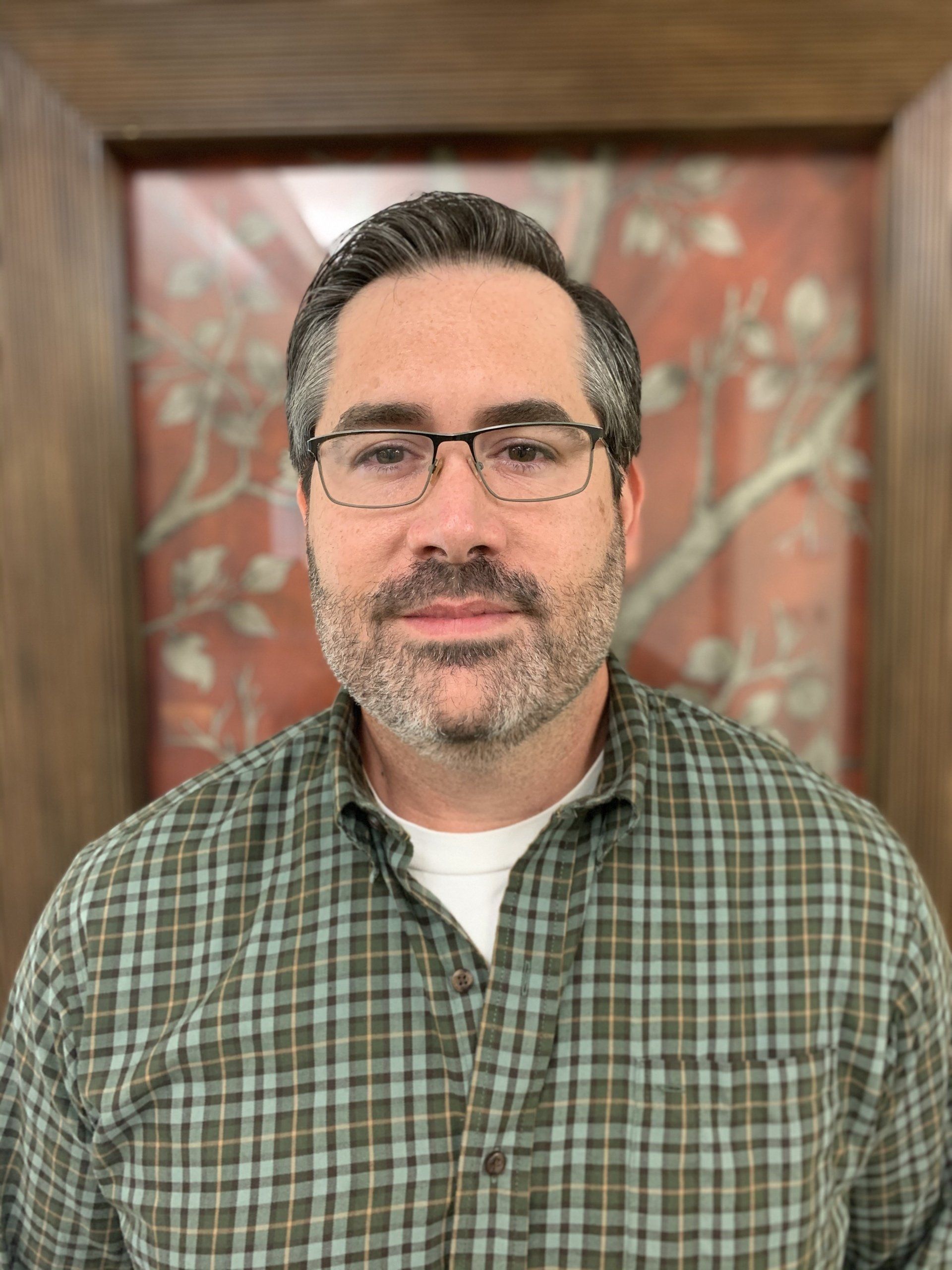 A man wearing glasses and a plaid shirt is standing in front of a picture.