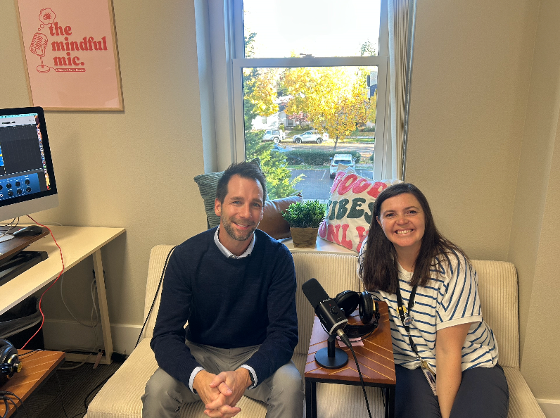 Two people sit on a couch in front of a window, podcasting. A microphone is on a table between them.