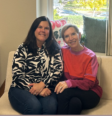 Two women seated on a couch, smiling. One wears a zebra-print jacket, the other a pink and orange shirt. Window in background.