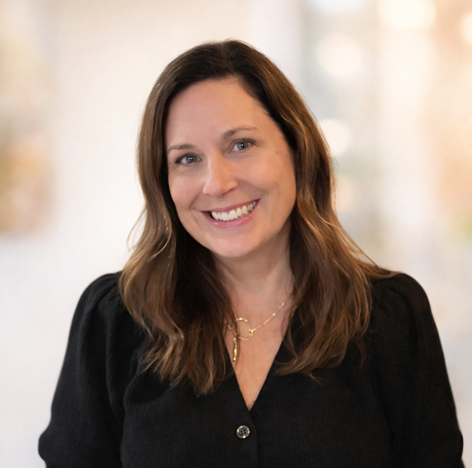 Woman with shoulder-length brown hair, smiling, wearing a brown sweater and a gold necklace, outdoors with blurred background.